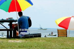 man sitting at a picnic table under a rainbow umbrella, playing the guitar overlooking Rock Hall Beach with boats on the horizon